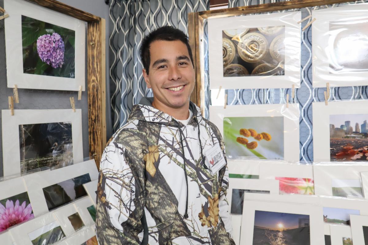 Man smiles posing in front of matted photographs for sale