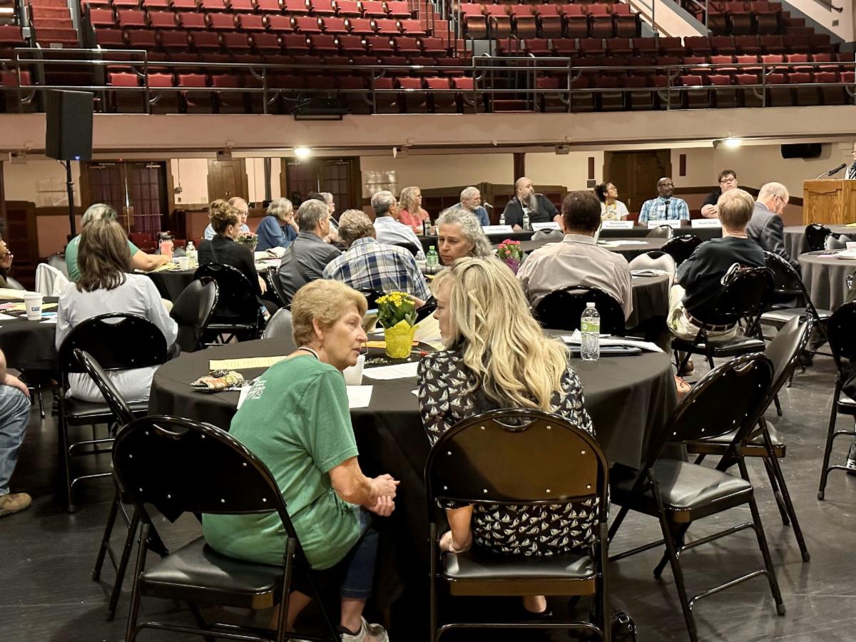 Large group of people at tables set up on a stage having a meeting