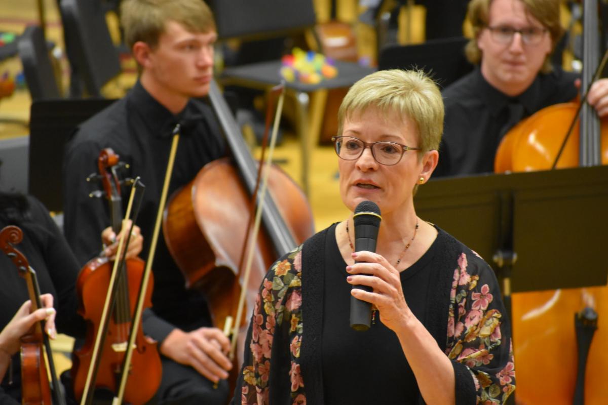 Melanie Loy with a microphone in front of high school orchestra