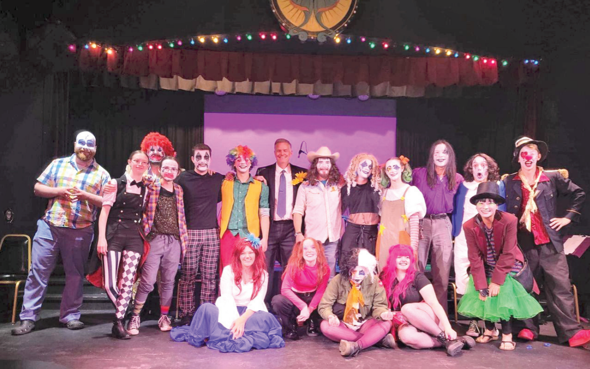Cast of a play on a stage, most dressed in clown clothes