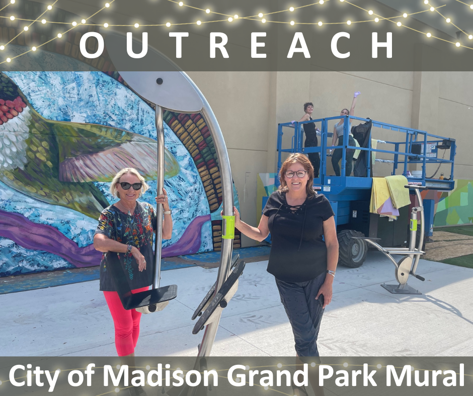 2 people pose in front of a partially complete outdoor mural of a humming bird while two other artists are waving from a crane to continue working on the mural