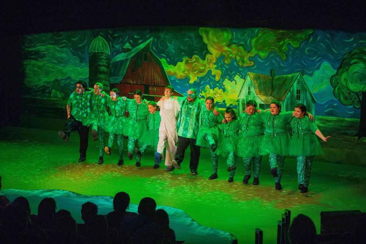 Stage lit in green backdrop of stylized country scene and group of dancers on stage