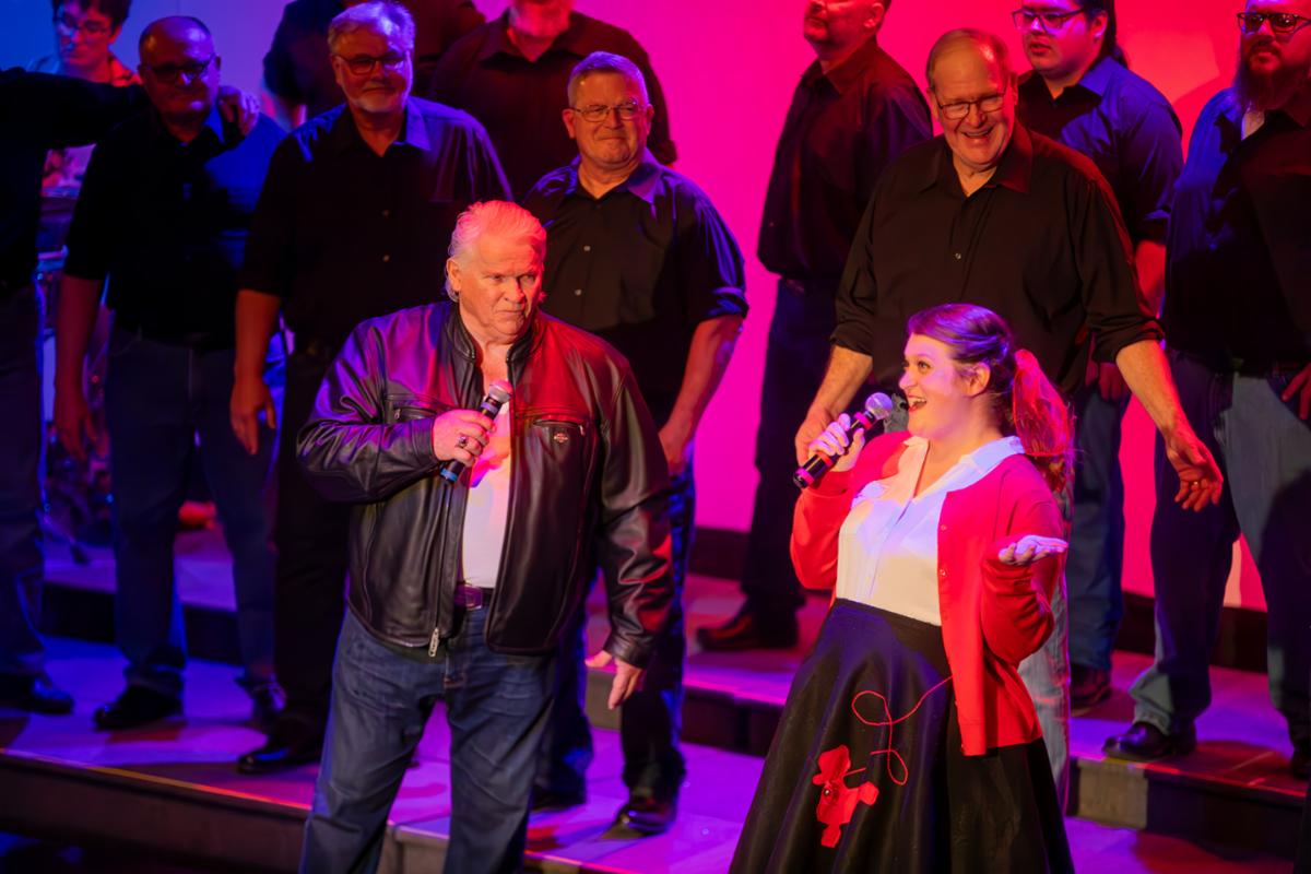 A group of singers on stage, the two main singers up front are a man dressed in a leather jacket and a woman in a poodle skirt