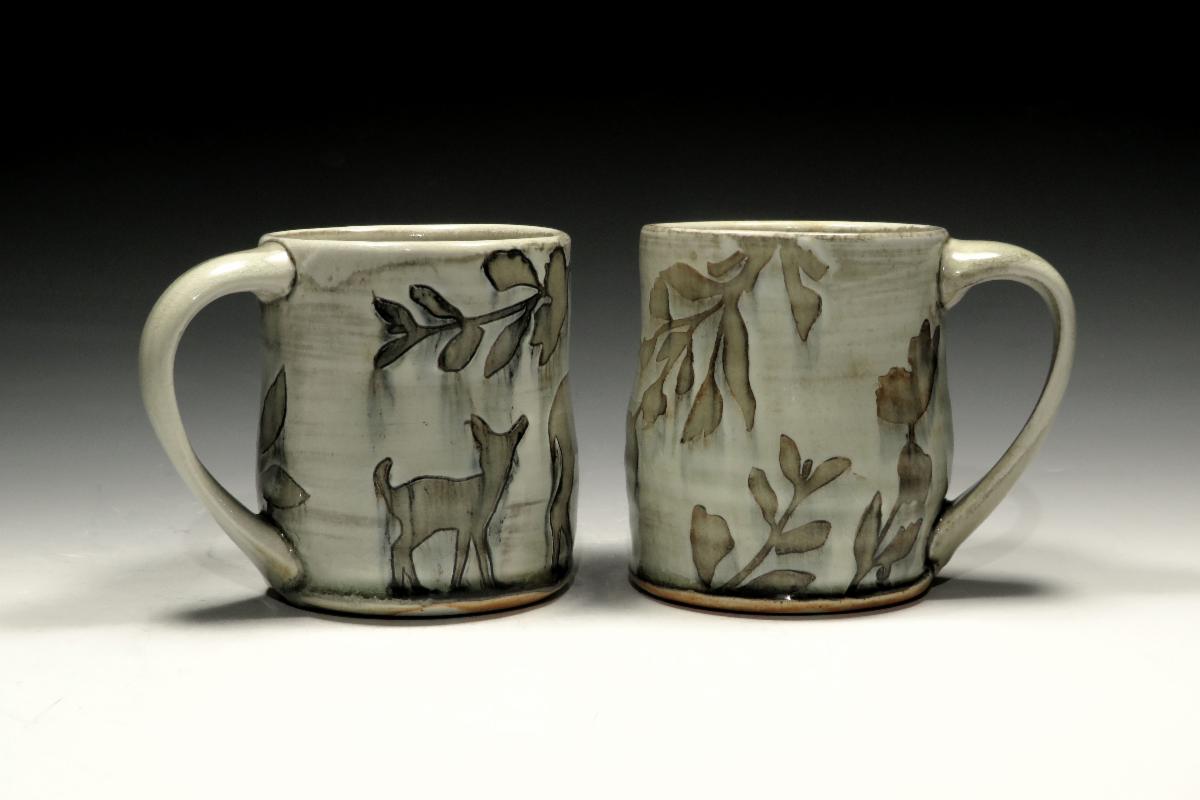 Two light colored mugs with leaves and deer motifs