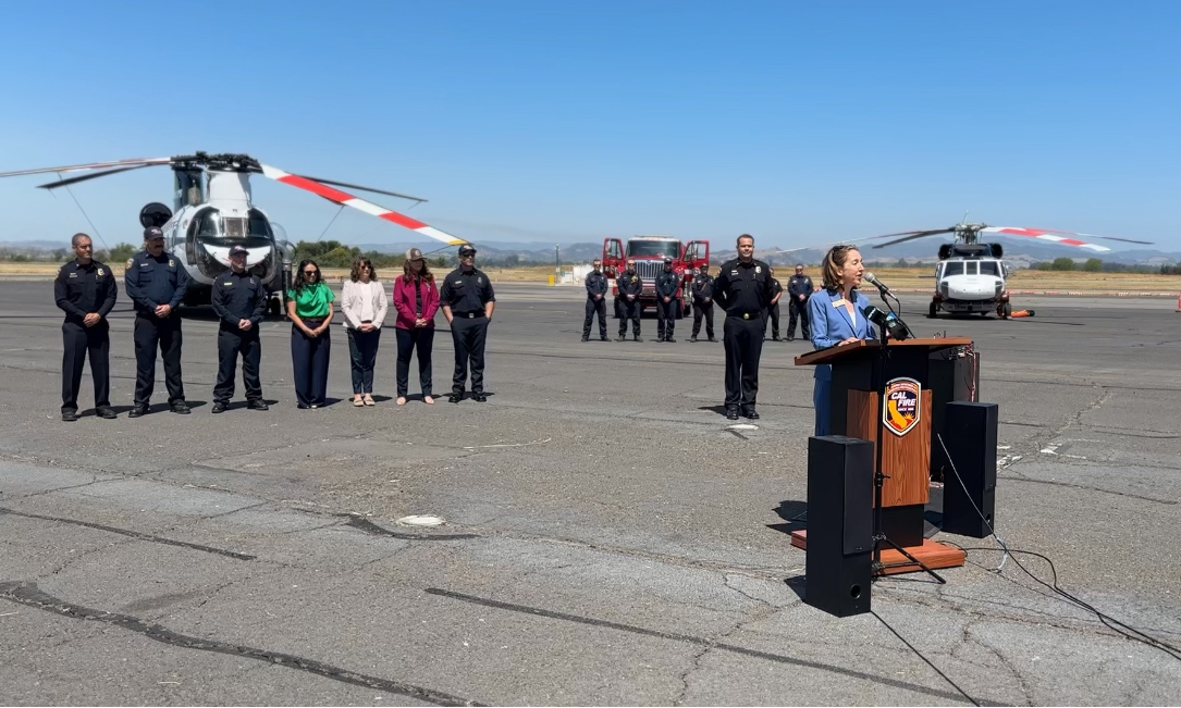 Anne Cottrell speaking at podium during CAL FIRE press conference with helicopter fire personnel and fellow members of the Board of Supervisors behind her