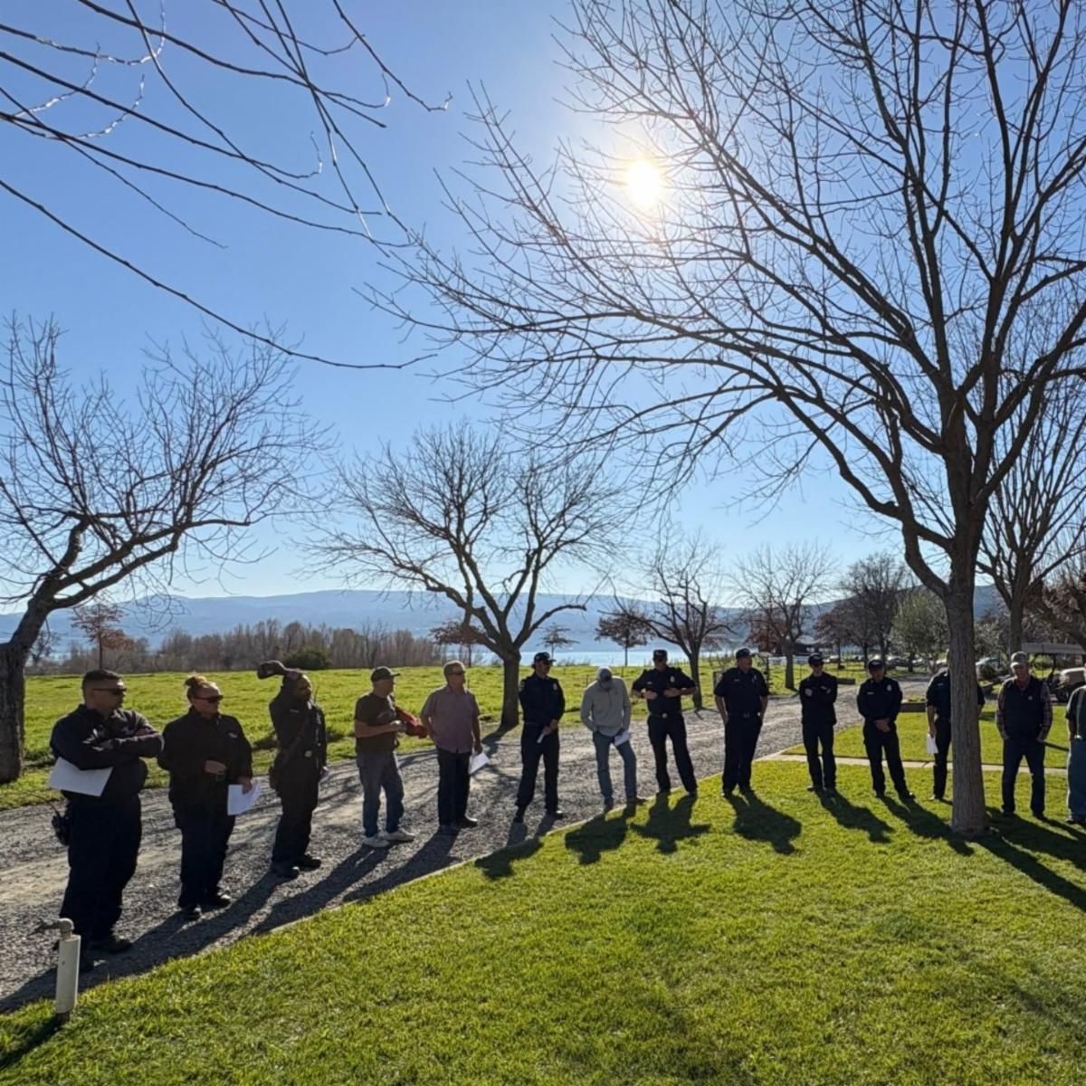 A group of firefighters and members of the Berryessa community stand together outside in a half circle.  The sun shines overhead with green grass beneath their feet.