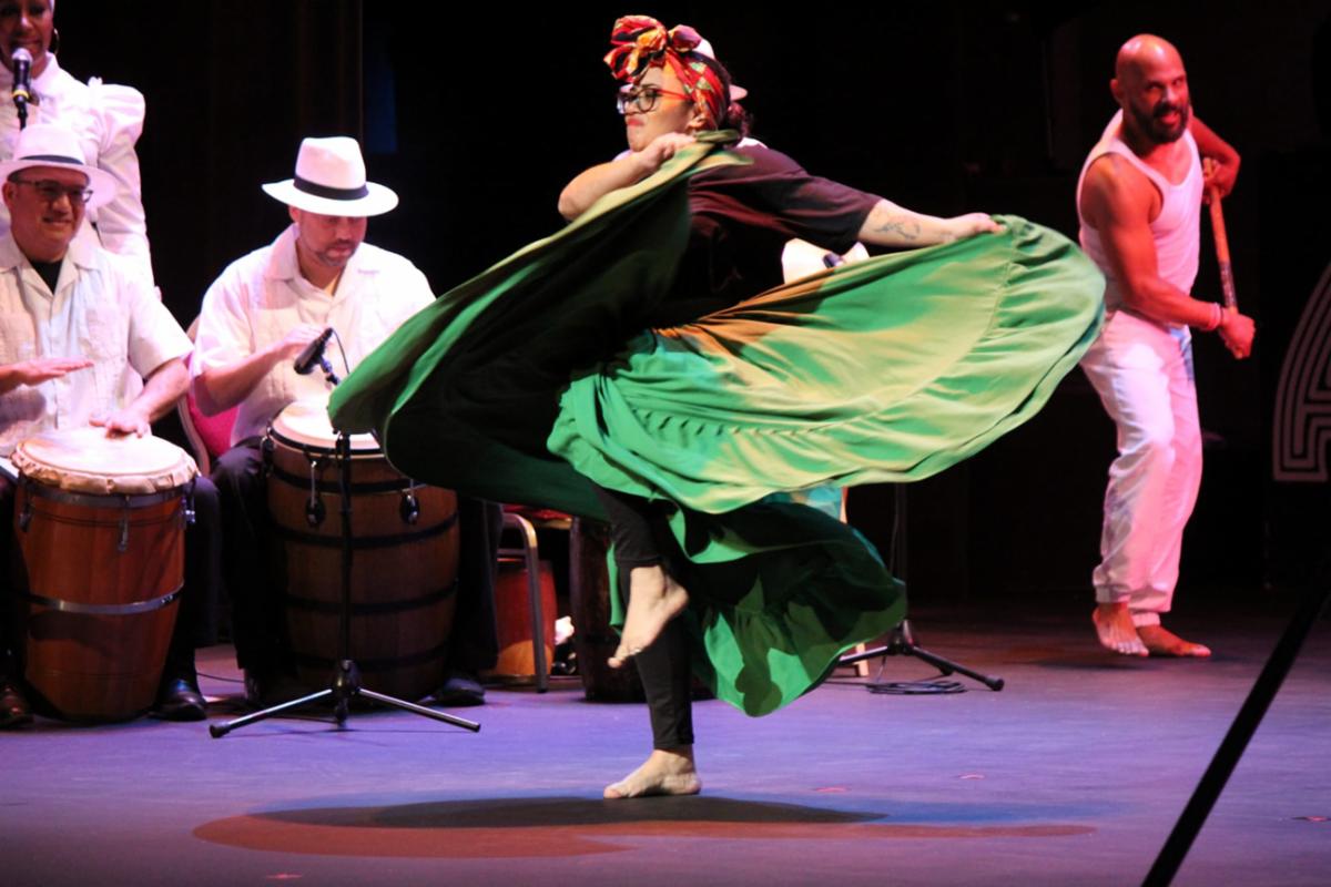 Featured front and center is an hispanic female dancer on stage with drummers and male dancer in the background