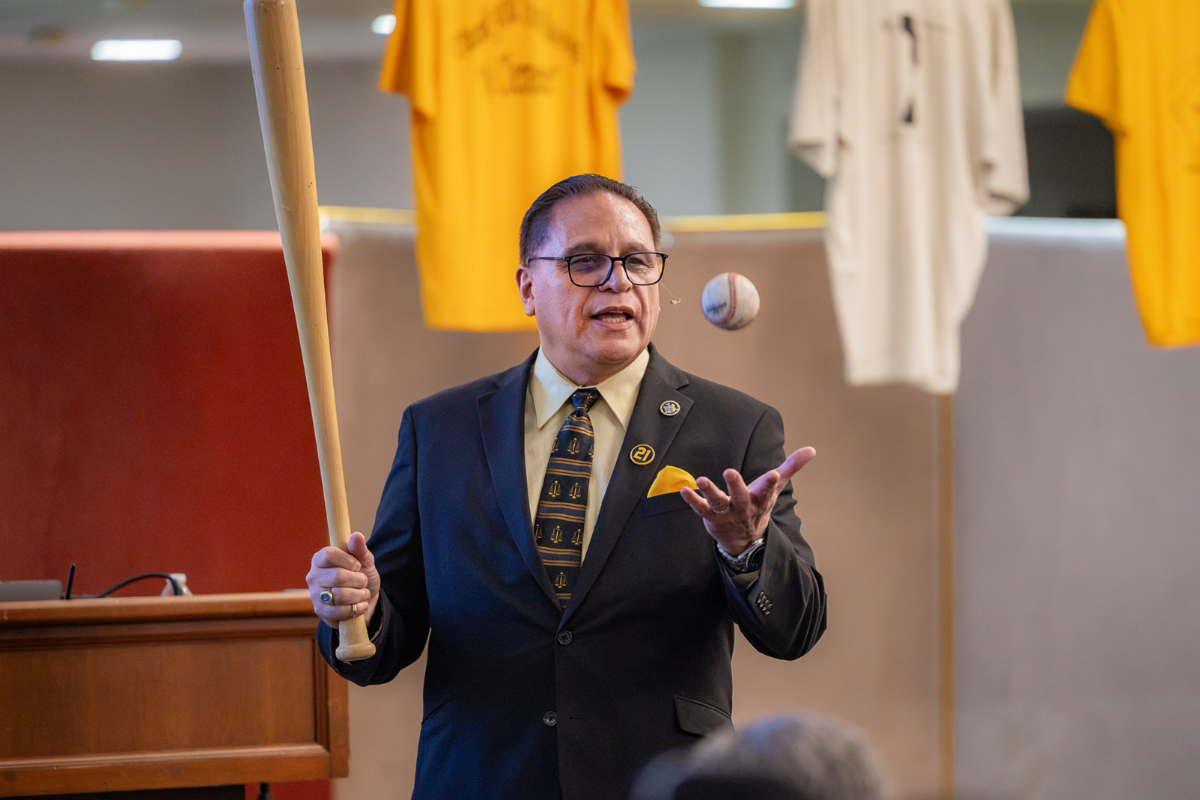 Hon. Eliezer Rodriguez, J.C.C., founder of Project Club Clemente Inc holds a wooden bat while gesturing toward a ball suspended in midair during a presentation