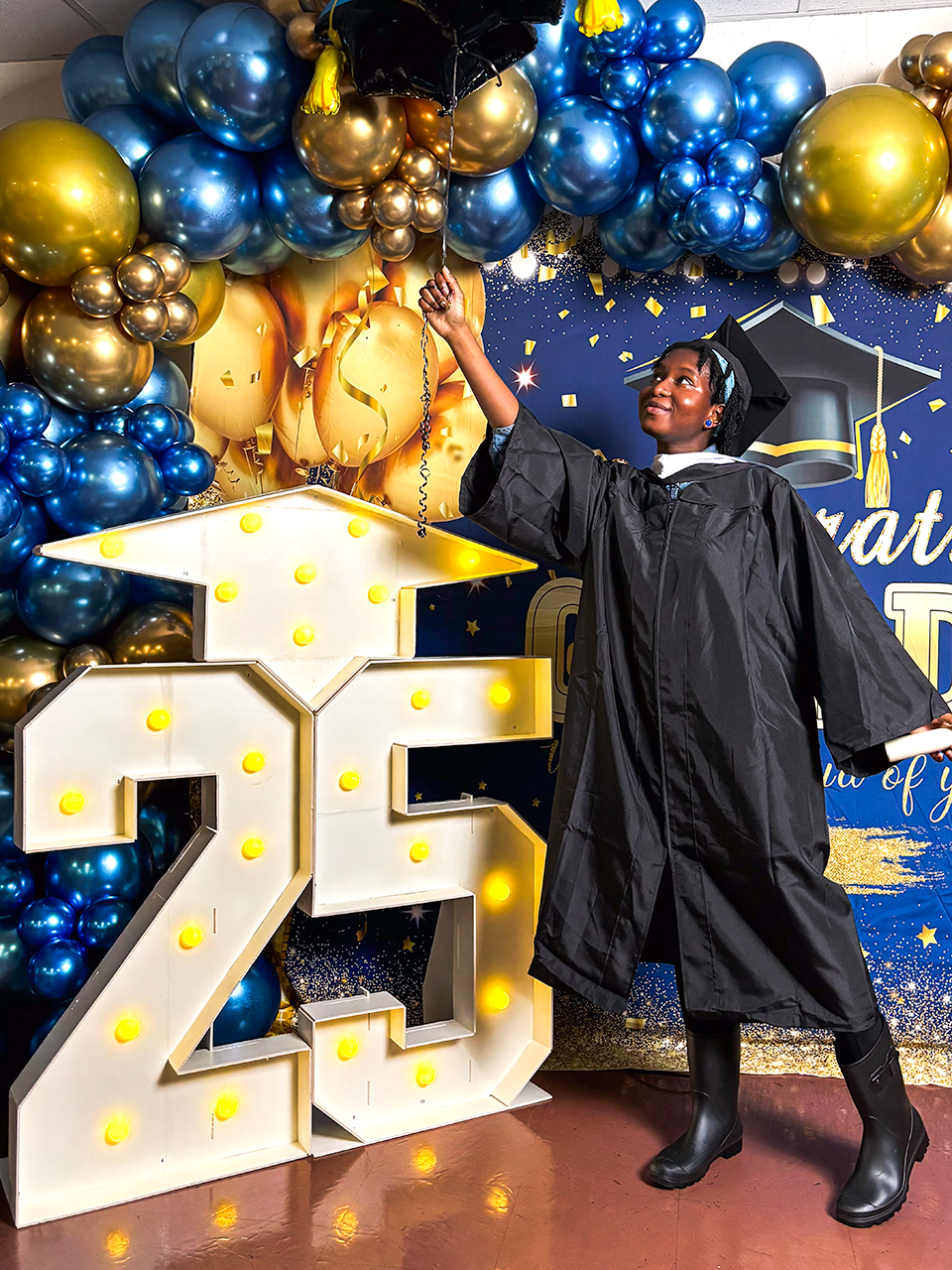 Student in graduation gown posing beside the number 25 and reaching for a balloon