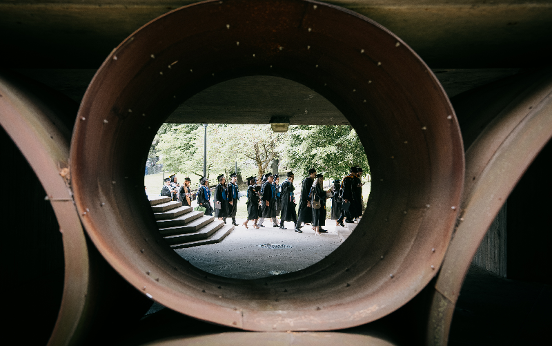 Graduates in caps and gowns walk across campus, framed through the circular opening of a large metal sculpture.