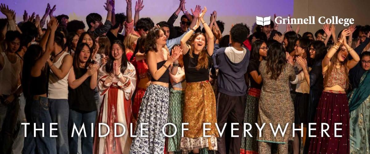 Students in multicultural attire gather on stage with their hands in the air.