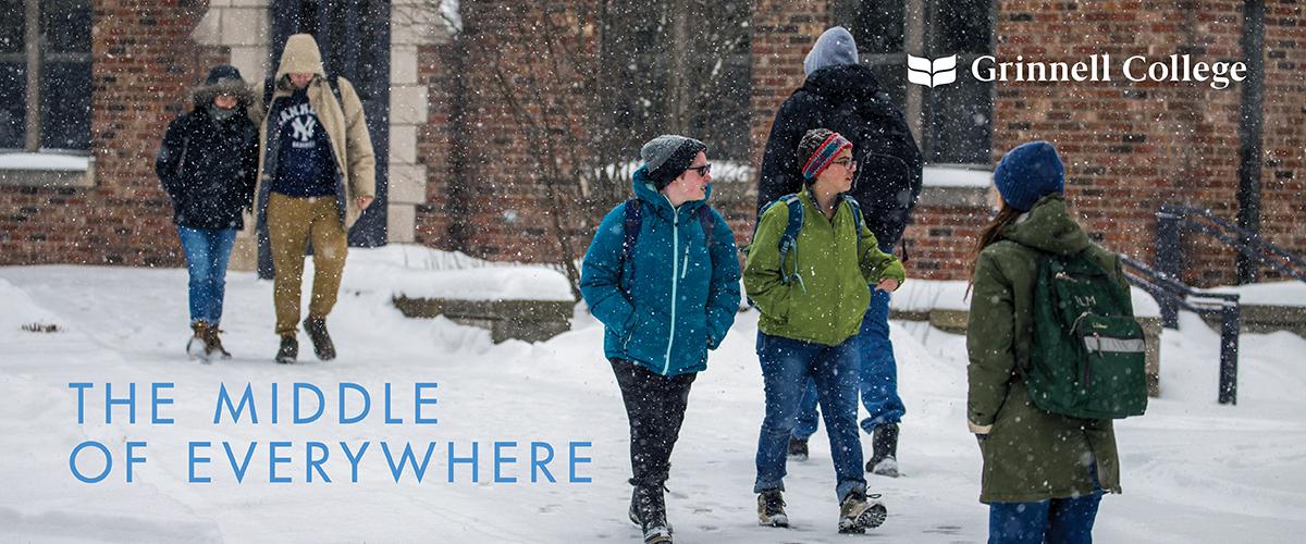 Students walk down a snowy sidewalk with snow falling in front of a brick building.
