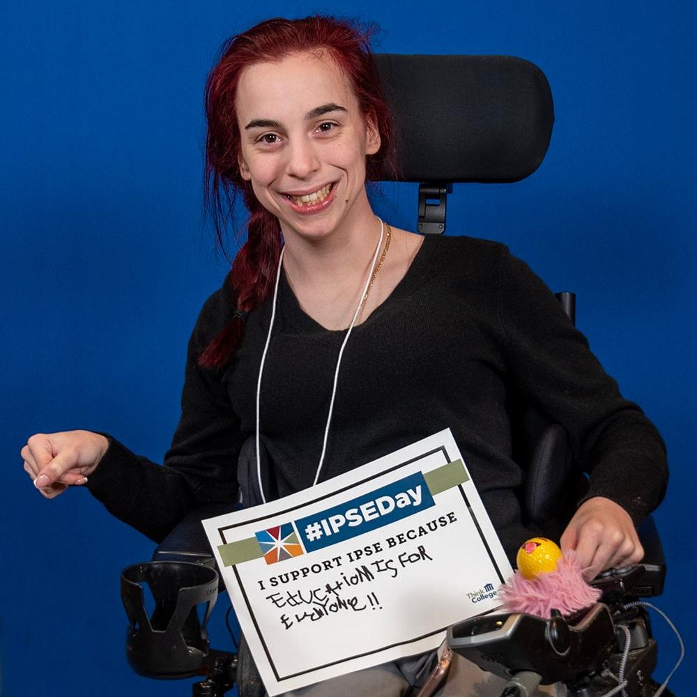 A person with red hair is sitting in a black motorized wheelchair against a blue background she is holding up a sign that says I support IPSE because Education is for everyone The person is smiling and wearing a black shirt and light colored pants