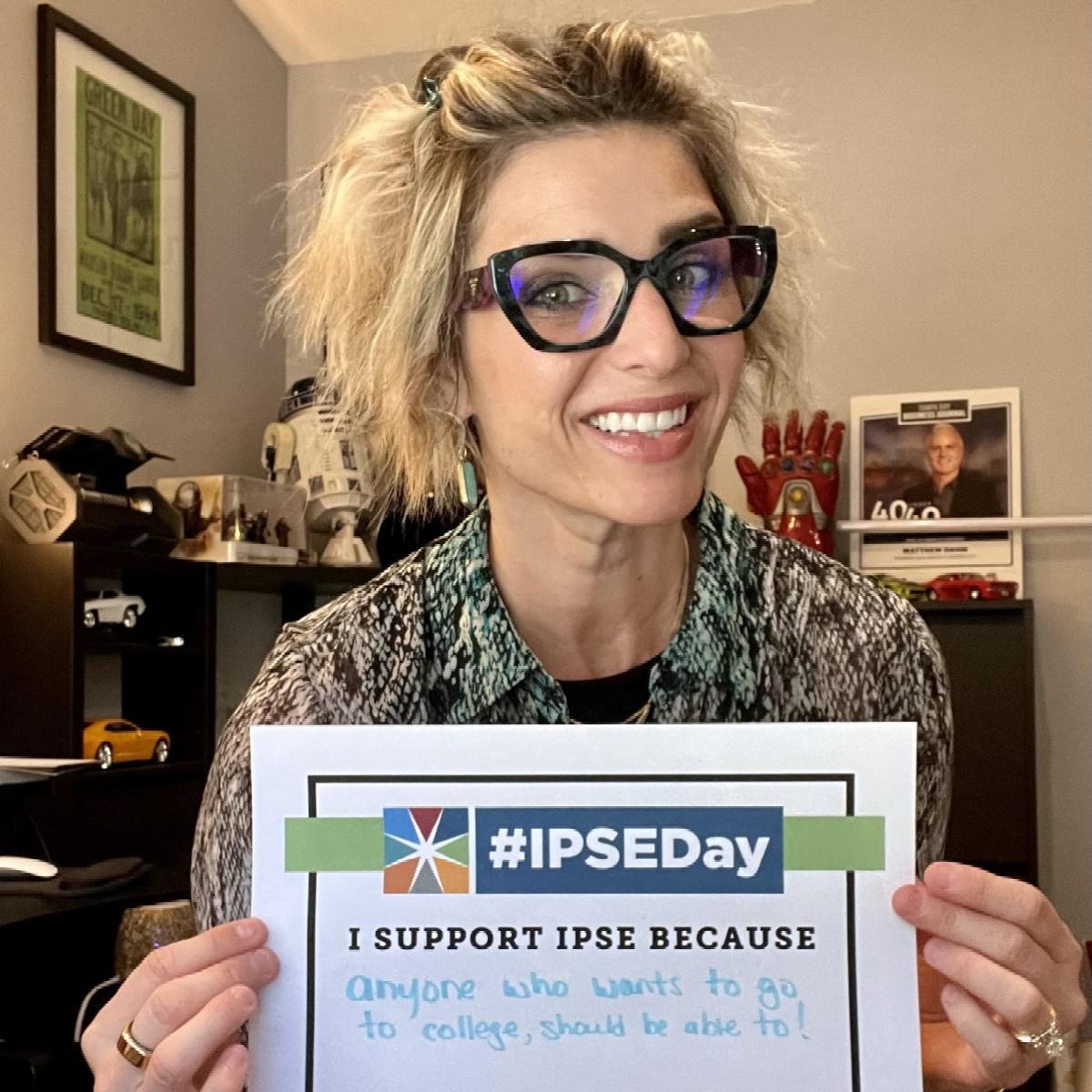 A person with short hair and glasses is holding a sign that reads IPSEDay I support IPSE because anyone who wants to go to college should be able to In the background there are shelves with various items including books and a toy car