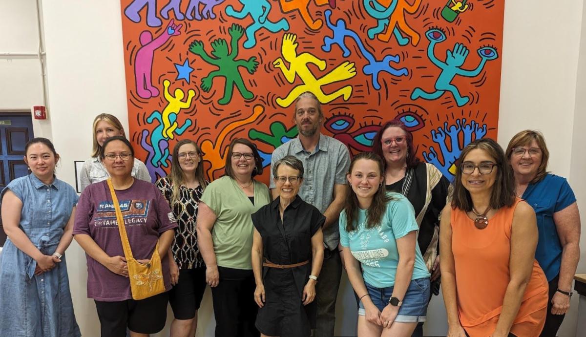 A group of twelve people stand smiling in front of a colorfully painted mural featuring abstract figures and bright vivid patterns and the diverse group is casually dressed