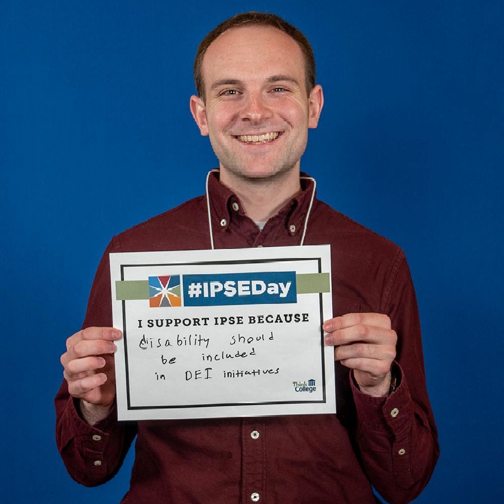 A person is smiling and holding a sign in front of a blue background The sign reads IPSEDay I support IPSE because disability should be included in DEI initiatives The person is wearing a maroon shir
