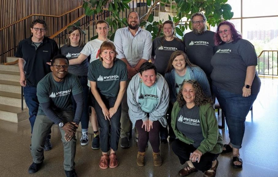 A group of twelve smiling people some wearing matching grey tee shirts standing together in front of a staircase with a large plant