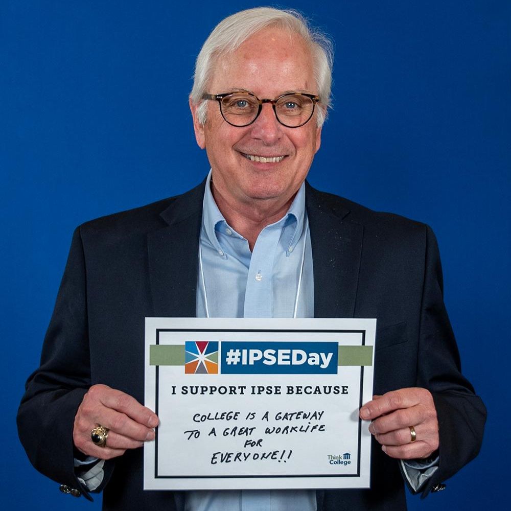 A man with white hair and glasses is smiling while holding a sign that reads I support IPSE because college is a gateway to a great work life for everyone The sign also has the hashtag IPSEDay at the top He is wearing a dark suit with a white shirt