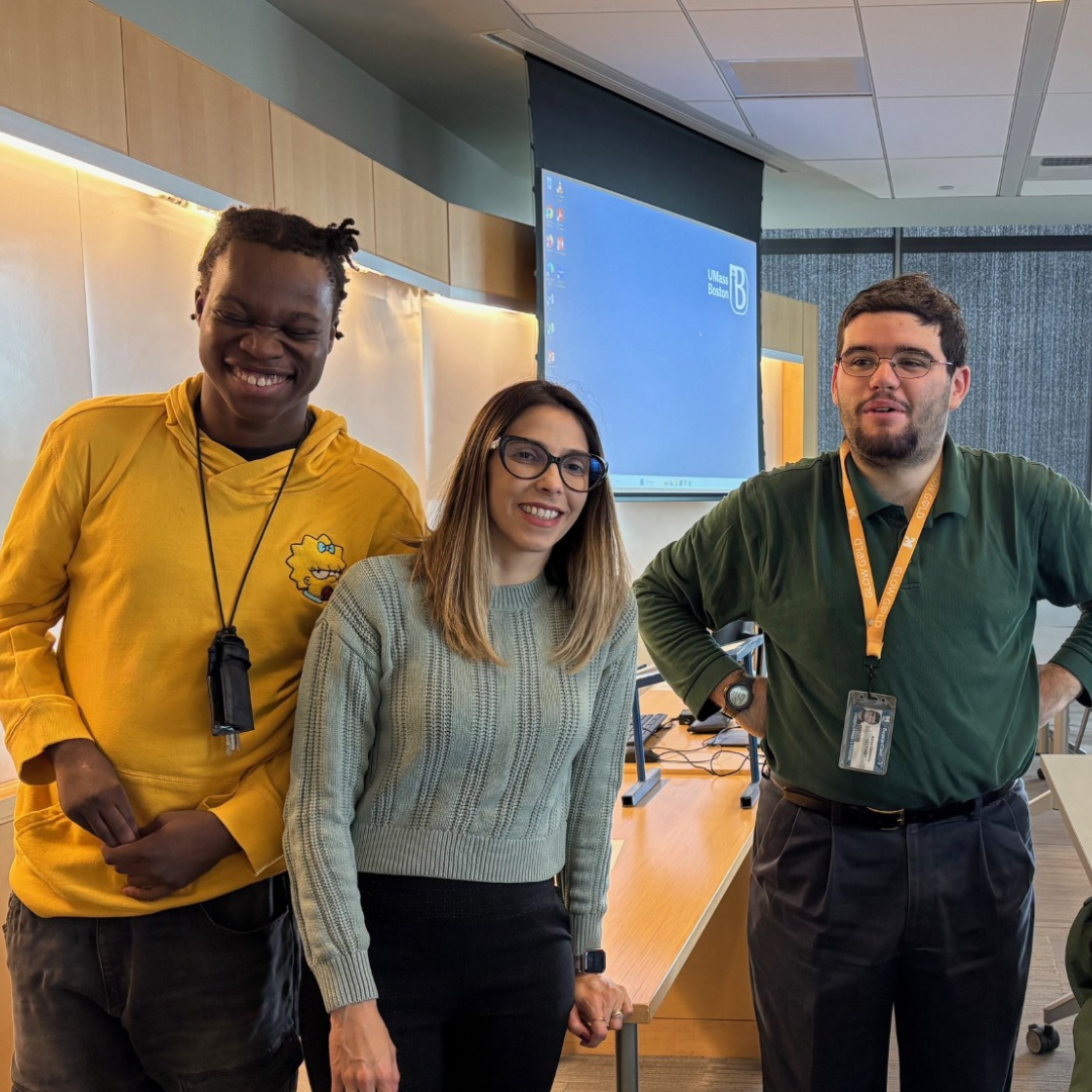 Three people stand in a conference room with a large table and chairs One wears a yellow shirt another a gray sweater and the third a green shirt They are smiling and the room has a projector screen and large windows