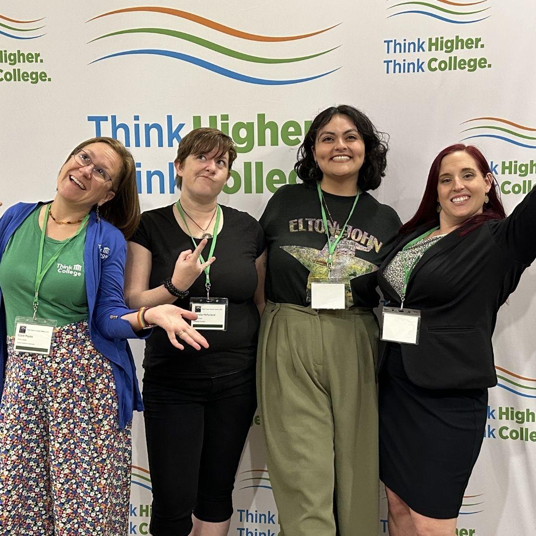 Four individuals two wearing green lanyards pose smiling in front of a backdrop with the words Think Higher Think College One wears glasses and a floral skirt another a black shirt another a green Elton John shirt and the fourth a black blazer and