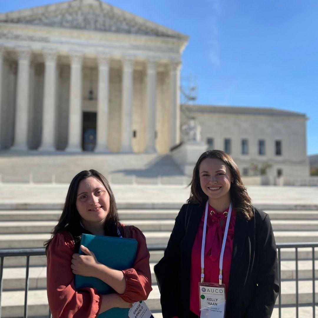 Two individuals pose in front of a federal building with columns in Washington DC One holds a folder and both wear ID badges The sky is clear and blue