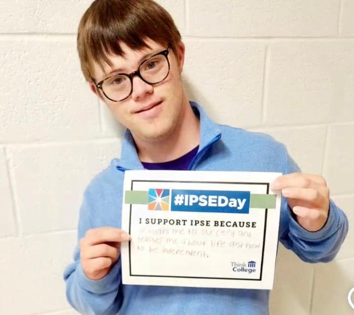 Young person with intellectual disability holding a handwritten sign with the IPSEDay logo that reads I support IPSE Day because It helps me to succeed and teaches me about life and how to be independent. 