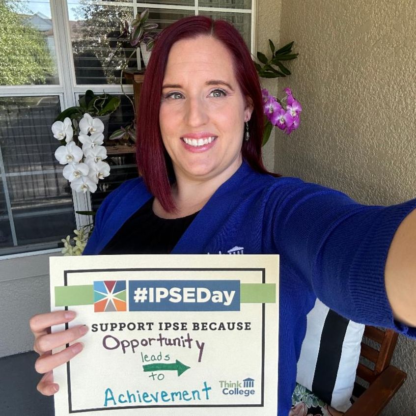 A person with shoulder length red hair wearing a blue sweater is holding a sign that reads IPSEDay SUPPORT IPSE BECAUSE Opportunity leads to Achievement Think College They are standing on a porch with a window and plants in the background