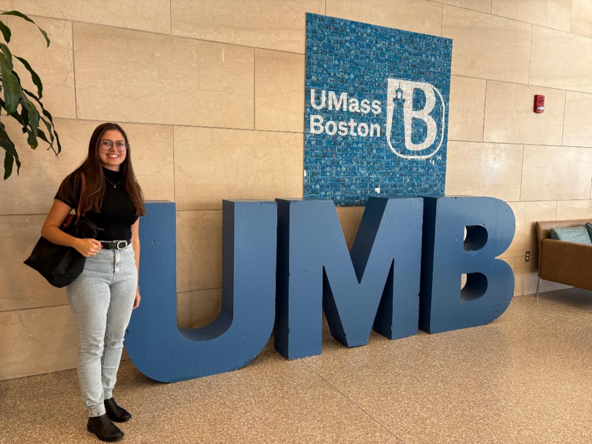 A person wearing a black shirt and jeans stands smiling next to large blue letters UMB inside a building A UMass Boston logo is displayed on the wall above