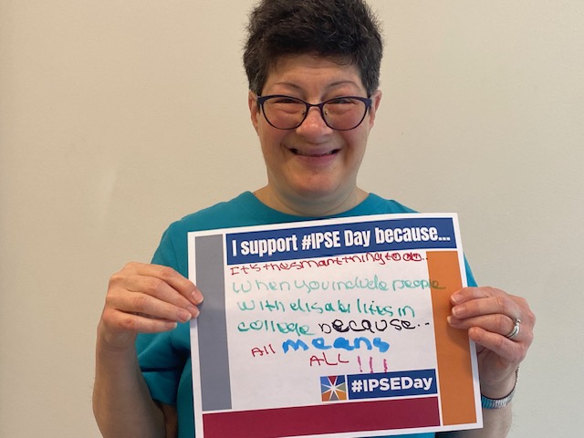 White woman which short dark hair and glasses wearing blue shirt holding up a sign with the IPSE Day logo and the sign reads I support IPSE day because it is the smart thing to do when you include people with disabilities in college because all