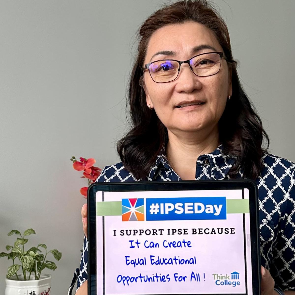 A person with glasses and a blue patterned shirt holds a sign that reads I support IPSE because it can create equal educational opportunities for all Think College with the hashtag IPSEDay at the top Plants are visible in the background