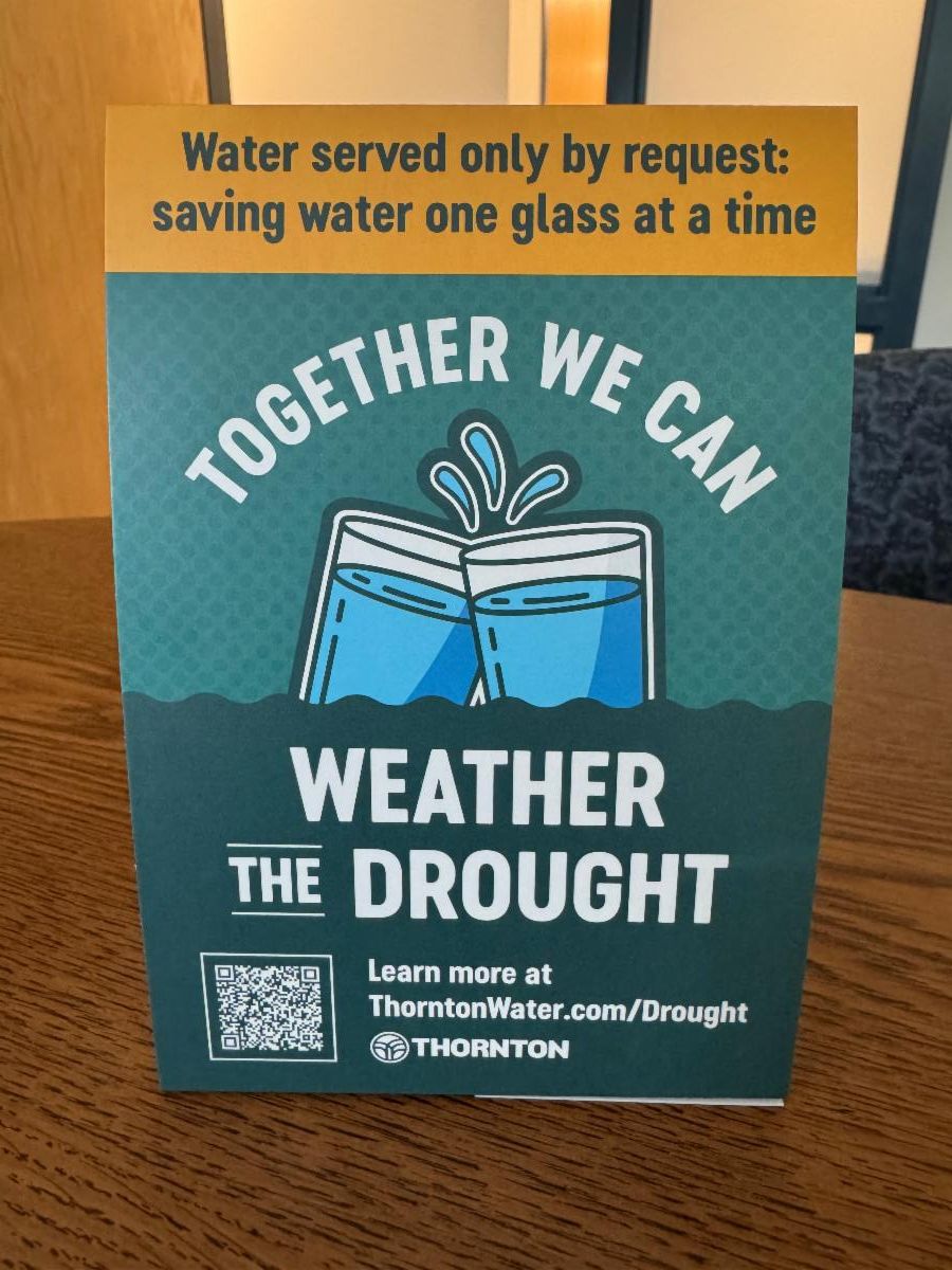 A teal and yellow table tent sign with a graphic of two clinking glasses of water. Text reads: "Water served only by request: saving water one glass at a time. Together we can weather the drought." At the bottom, a QR code sits next to text that reads: "Learn more at ThorntonWater.com/Drought" above the Thornton logo.