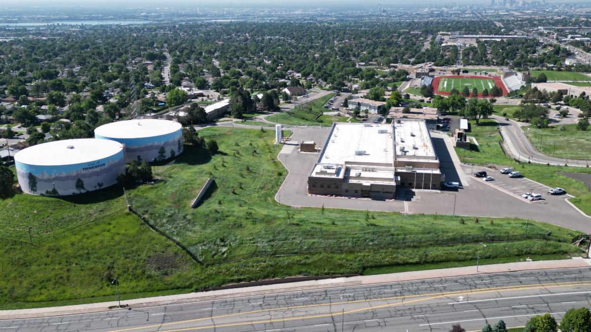 Aerial photo of a large building on a green hill, with two large cylindrical water tanks on the left, with trees and a football field in the distance. 