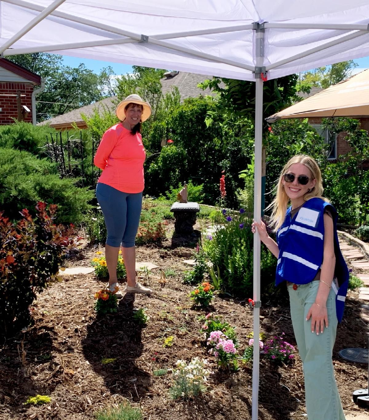 Two women smiling under a white canopy tent in a garden filled with mulched beds and colorful flowers. One woman wears a sunhat and bright coral shirt, while the other wears sunglasses and a blue volunteer vest.