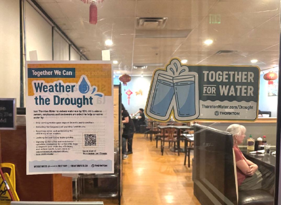 Water conservation signs reading "Together We Can Weather the Drought" and "Together For Water" posted on a restaurant's glass window.