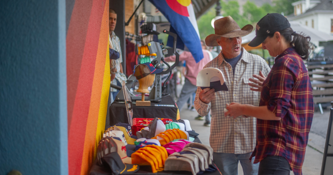 A person sells a hat to another person outside a shop in a mountain town