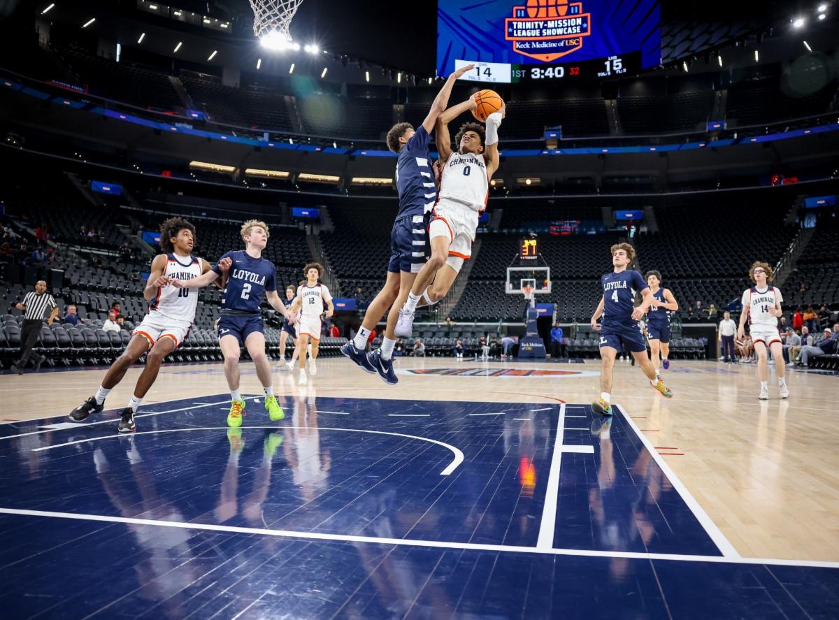 Male high school students playing basketball in an indoor gym with a player jumping mid-air while trying to throw the basketball in the hoop with a play defending him who is also mid-air. 