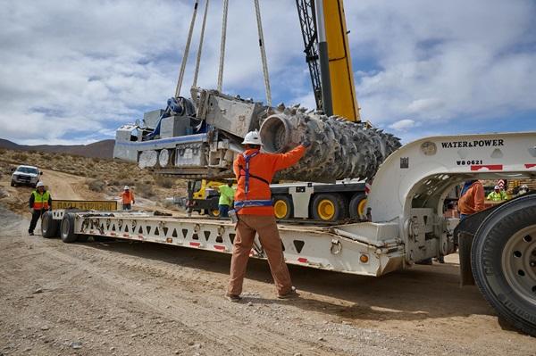Workers in safety gear help guide heavy equipment off a truck.