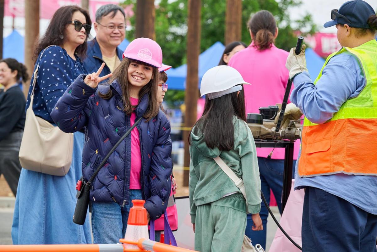 Girl in pink hard hat posing and smiling for a picture at the Women’s Expo event. Another girl in green with a white hard hat is next to her observing a job demonstration. 