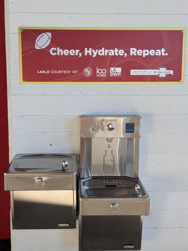 A dual-model silver metallic drinking fountain and modern reusable bottle filler affixed to a white concrete wall. Above them, a rectangular red sign with a gold border reads in white lettering, “Cheer, Hydrate, Repeat.” A football icon is above the text.