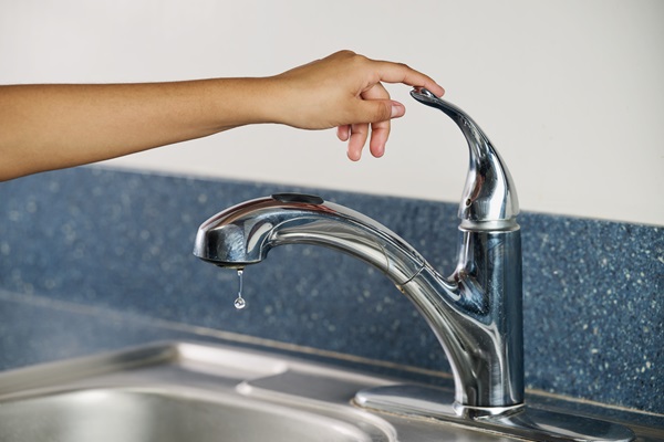 Hand of a person turning off the sink faucet with a water droplet dripping.