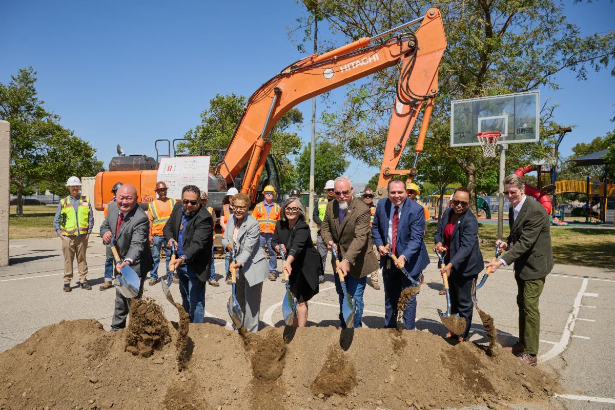 City leaders stand in a line holding shovels and lifting dirt during the David M. Gonzales Recreation Center groundbreaking event for the Stormwater Capture Parks Program. They are positioned in front of an orange excavator, with construction workers in safety vests and hard hats behind them. The scene takes place outdoors on a sunny day near a basketball court and a playground, with trees and park equipment visible in the background.