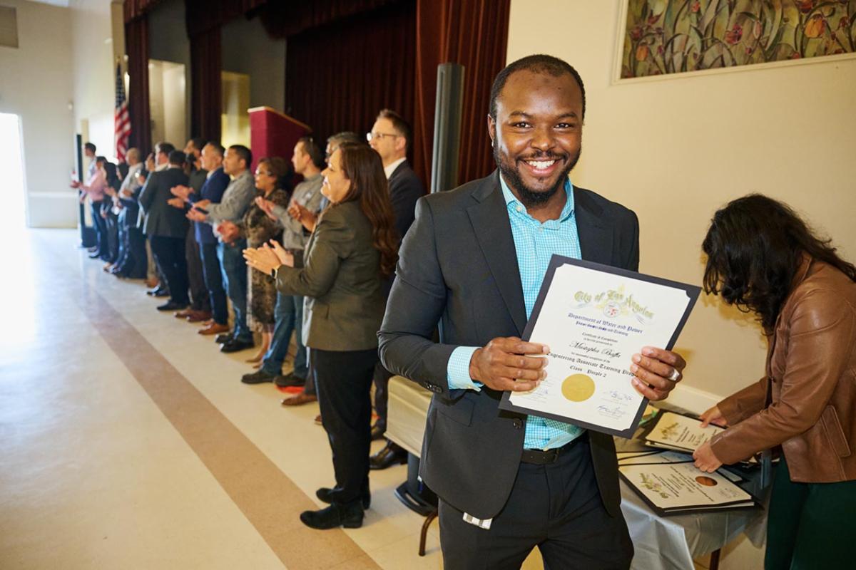 Man holding a certificate smiling and posing for a photo with a group of lined up people clapping behind him in an auditorium setting. 