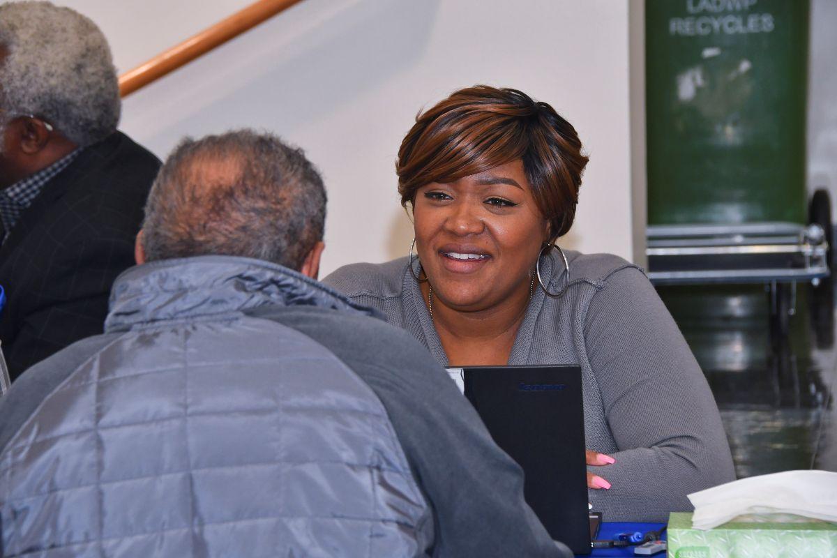 A woman with short black and brown hair a gray cardigan and hoop earrings smiling and sitting down with a laptop in front of her while looking at a man who is also sitting down. 