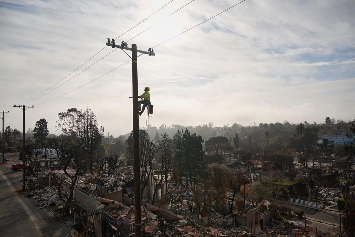 Line worker climbing an electric pole outdoors with various household debris on the ground level.