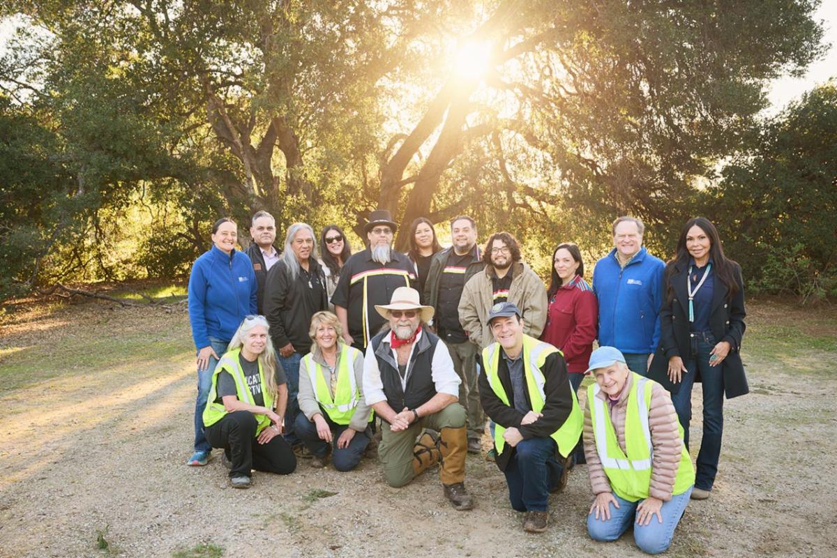 Group of individuals posing for a picture outdoors with some standing and some kneeling with trees and the sunrise in the background.