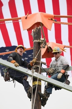 Two male line workers on a pole competing with a USA flag in the background. 