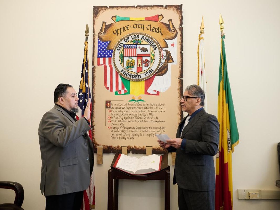 Man in gray suit holding up his hand and standing in front of another man in a black suit with the City of Los Angeles sign and flags in the background. 
