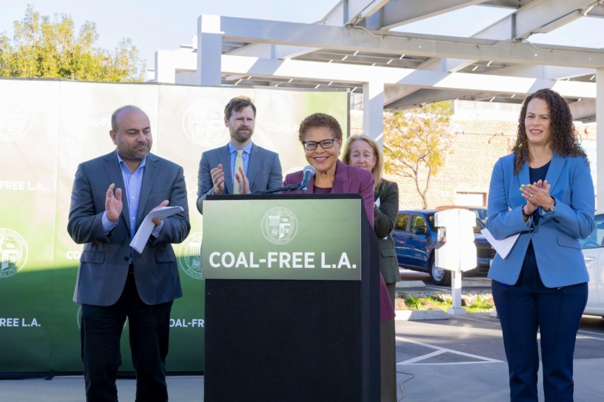 Smiling woman with glasses is standing at a podium with a microphone, while four people are in the background clapping.