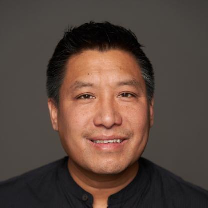 Smiling man in a black shirt is indoors with a dark gray background. 