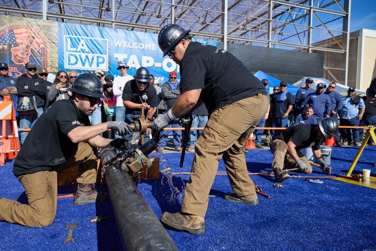 Two men wearing hard hats and gloves work together to fix a water pipe during a skills competition as spectators watch.